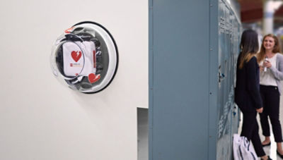 CR2 AED wall unit hangin beside two students leaning against lockers in school hallway