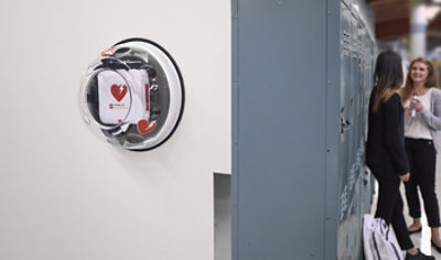 CR2 AED wall unit hangin beside two students leaning against lockers in school hallway