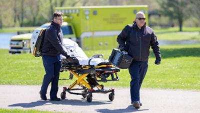 Two paramedics transport a patient on a stretcher toward an ambulance in a park setting