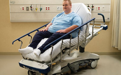 Pacjent na wózku transportowym z serii Prime firmy Stryker w ułatwiającej wstawanie pozycji krzesła kardiologicznego