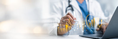 Healthcare professional using a stylus and laptop with an overlaid digital analytics dashboard showing charts, graphs, and healthcare data visualizations.