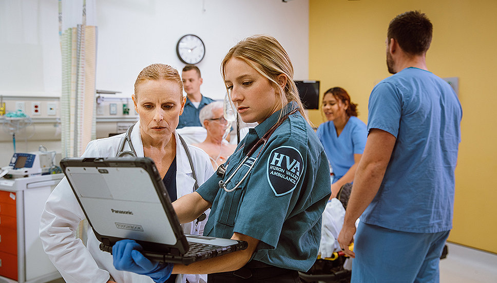 An EMT reviews patient data with a doctor while the patient and nurses on in the background