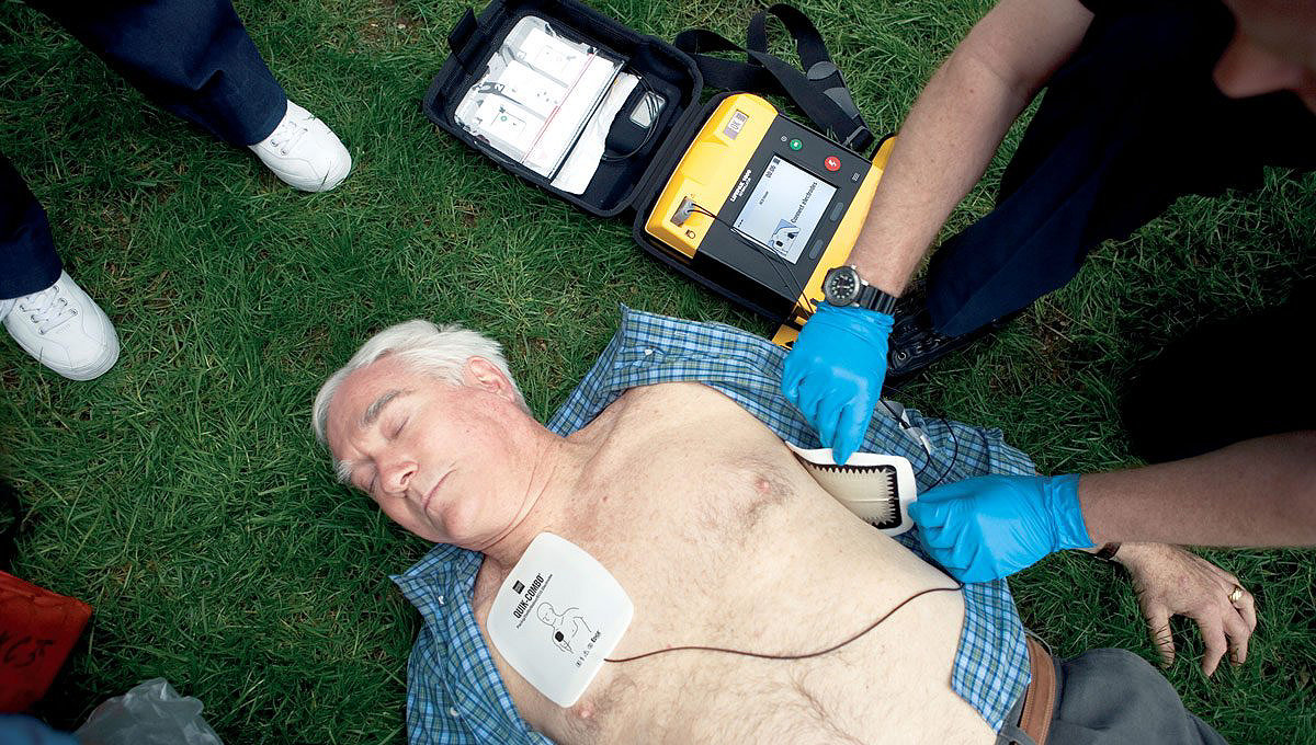 Bărbat întins pe sol, cu un defibrilator LIFEPAK 1000 conectat la torace.