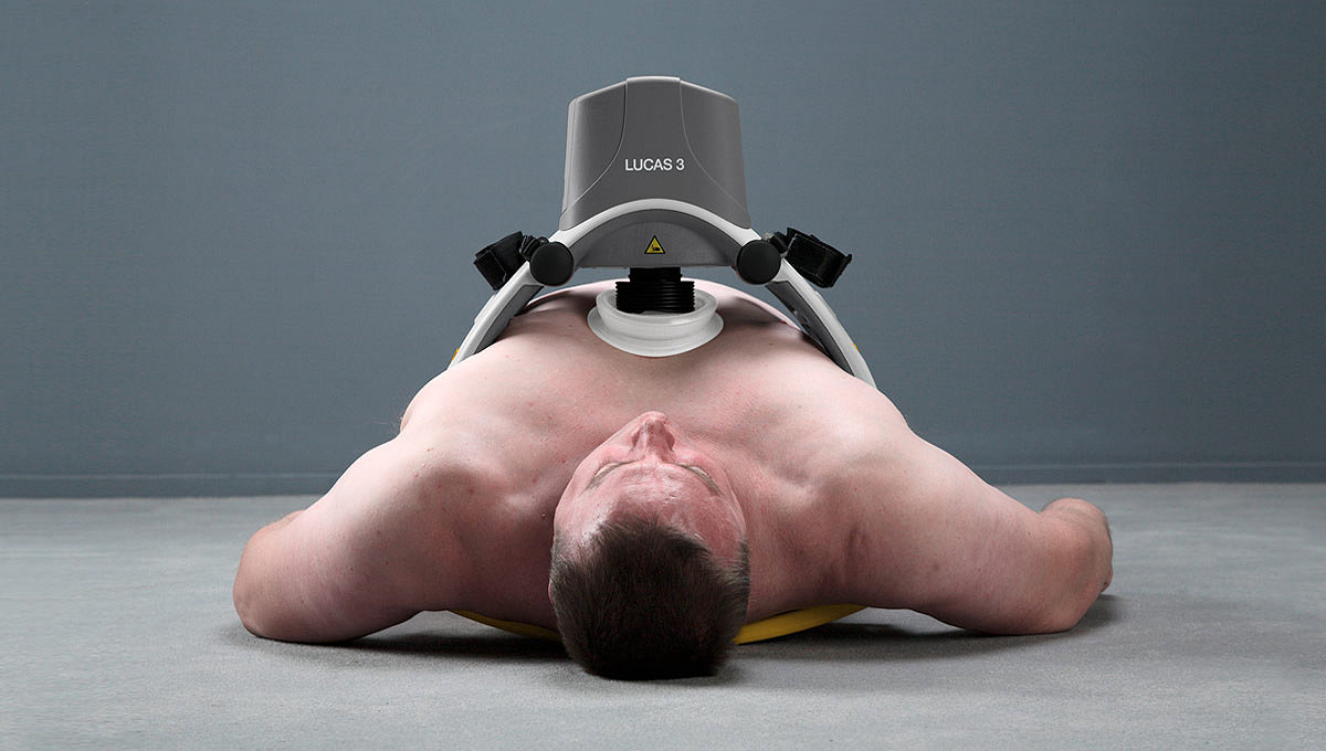 A large man laying on a floor demonstrates how the  LUCAS 3, v3.1 chest compression machine can fit larger adutls.