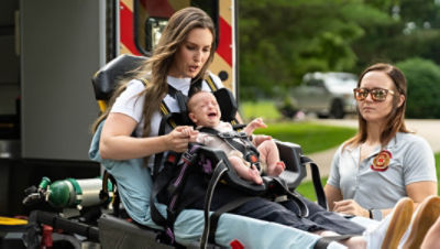 A mother riding the Power-PRO 2 stretcher toward an ambulance while comforting her crying baby that is in the Mini-PRO pediatric restraint
