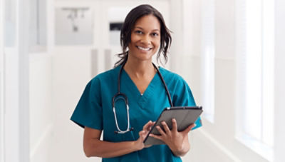 Nurse standing in hallway wearing blue scrubs holding tablet and smiling