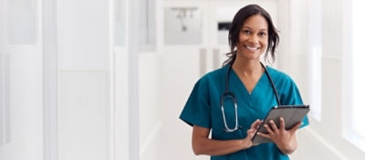 Nurse standing in hallway wearing blue scrubs holding tablet and smiling