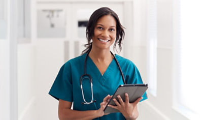 Nurse standing in hallway wearing blue scrubs holding tablet and smiling